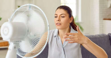 Woman suffering from summer heat and using electric fan to refresh in current of cool air. Young woman, tired of hot summer weather, sitting on sofa at home and enjoying cold air blowing in her face